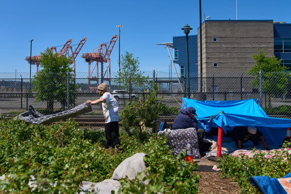 Three people are at a homeless encampment near a fence, industrial buildings, and cranes.