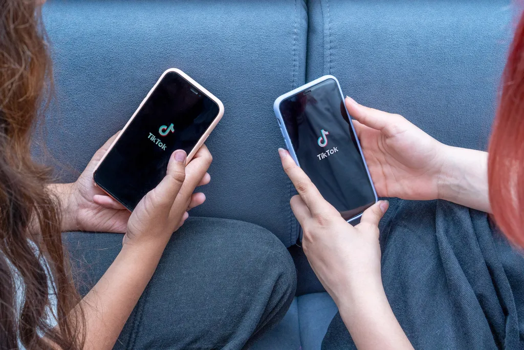 Two teenage girls sitting on a bed holding phones with the TikTok app open on their screens.