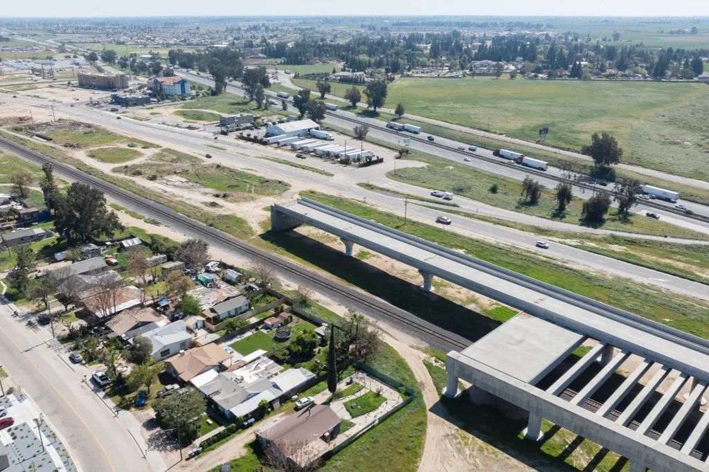 Aerial view of a highway running parallel to a railroad track, with residential areas and construction of a new overpass.