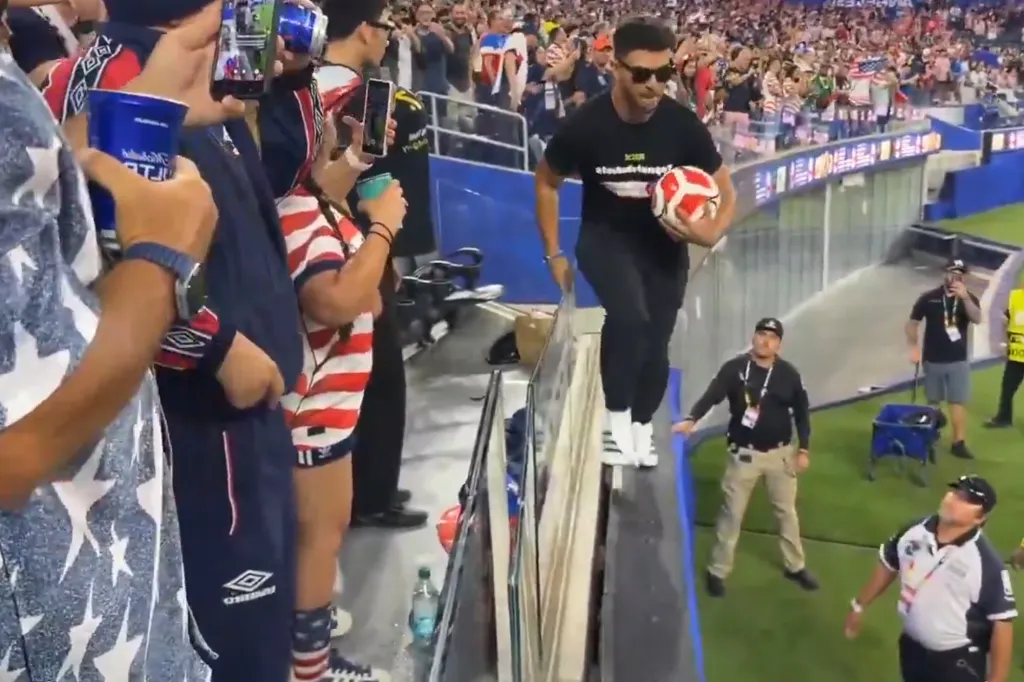 Man in black shirt and sunglasses holding a soccer ball walking across the top of a barrier with security guards nearby.
