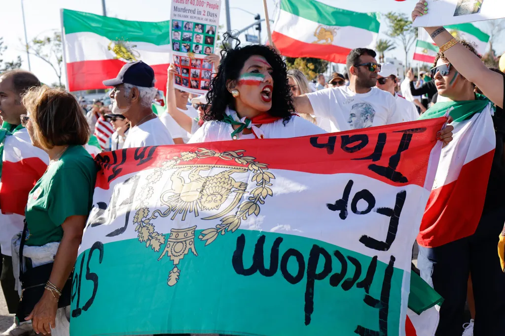 A woman with face paint in the colors of the Iranian flag holds a banner that reads "Freedom for Iran" while participating in a protest.