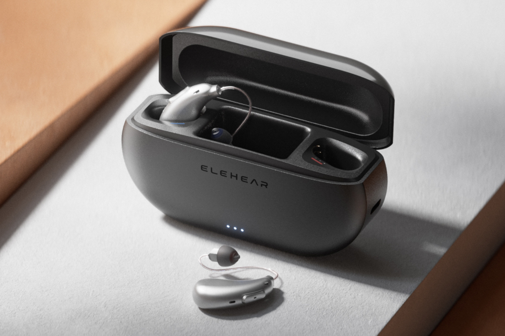 A silver hearing aid in a black charging case with another silver hearing aid next to it.