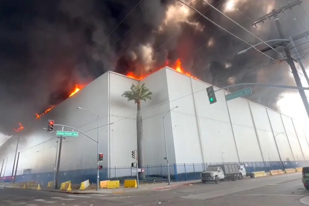 A storage facility in Boyle Heights on fire with flames visible on the roof and large plumes of black smoke filling the sky.