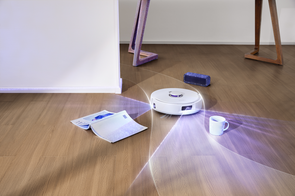 Robot vacuum with blue light showing its obstacle avoidance in a room with a magazine, speaker, and mug on the floor.