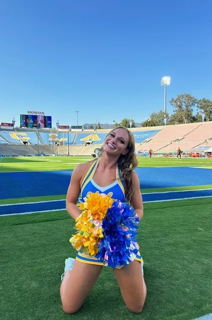 UCLA freshman Tenley Hill, dressed in a cheerleading uniform, smiles with pompoms on a football field.
