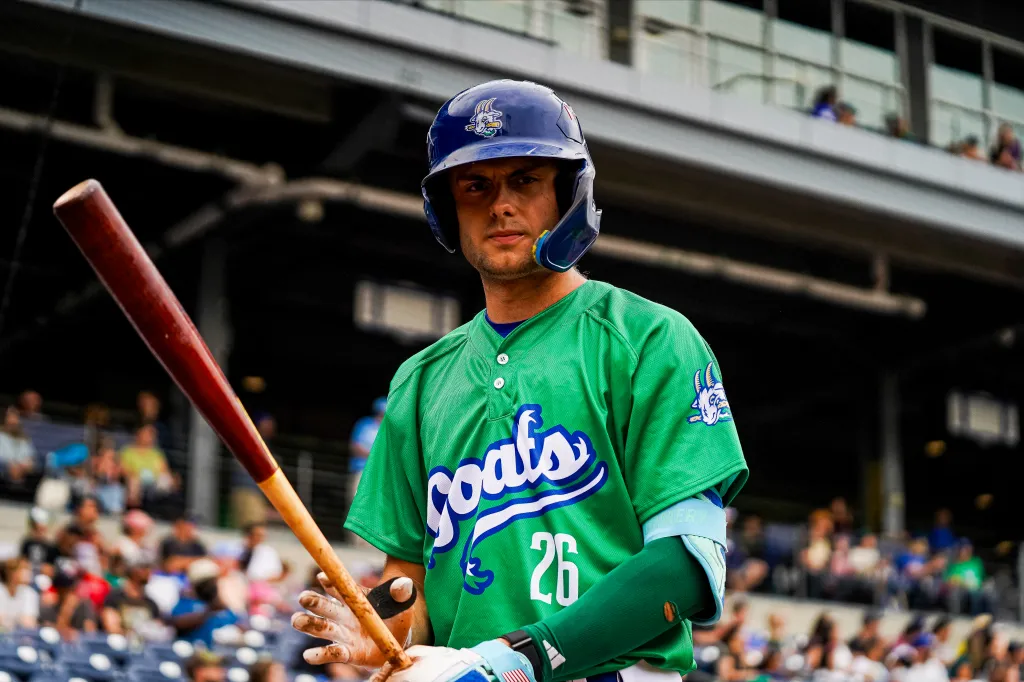 A baseball player in a green "Goats" jersey and blue helmet holds a bat, looking toward the camera.