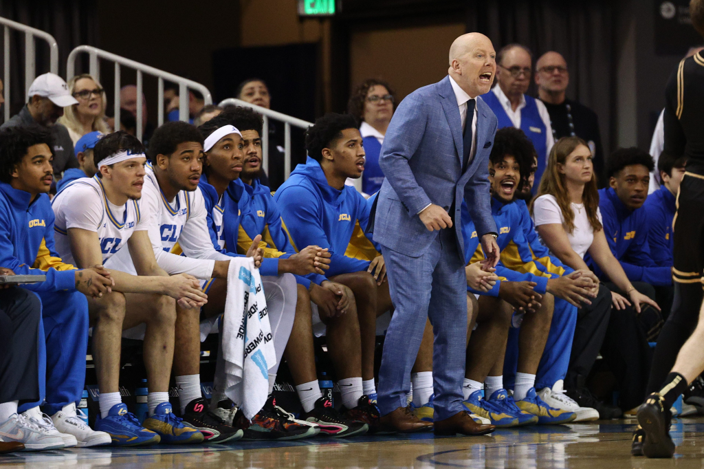 A basketball coach in a blue suit shouts from the sidelines next to seated players.