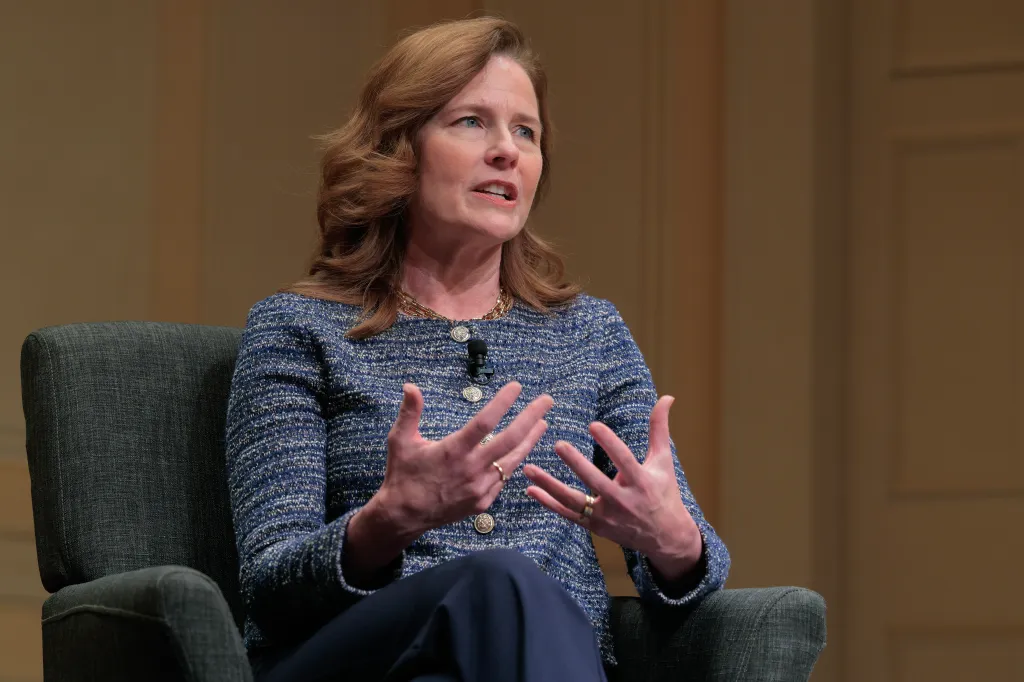 Amy Coney Barrett, in a blue tweed jacket with a gold necklace, sits in a gray chair, gesturing with her hands.