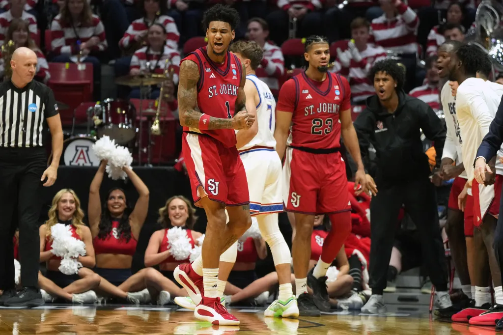 St. John's players in red and white uniforms on the basketball court during an NCAA game, with a referee and cheerleaders in the background.