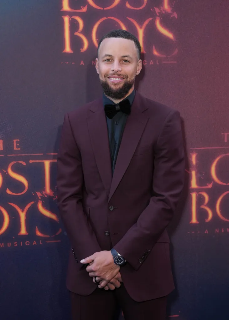 Stephen Curry in a burgundy suit and black bow tie, standing in front of a poster for "The Lost Boys: A New Musical."