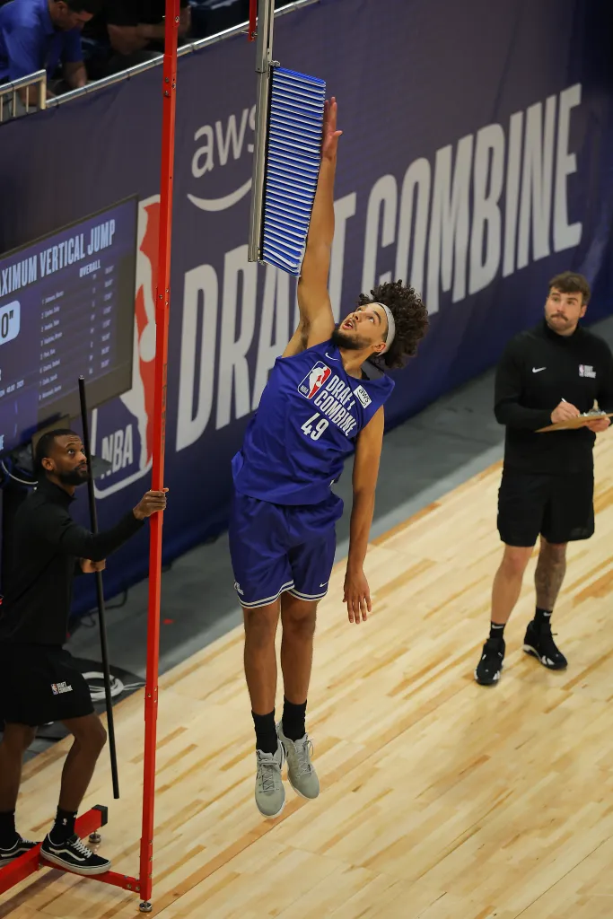 A basketball player in a blue jersey jumps to touch a vertical jump measurement apparatus.