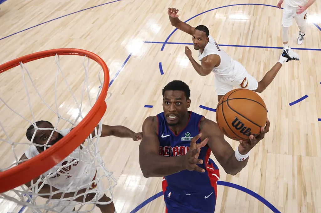 A basketball player in a blue and red jersey holding a basketball near the hoop, with two other players nearby on the court.