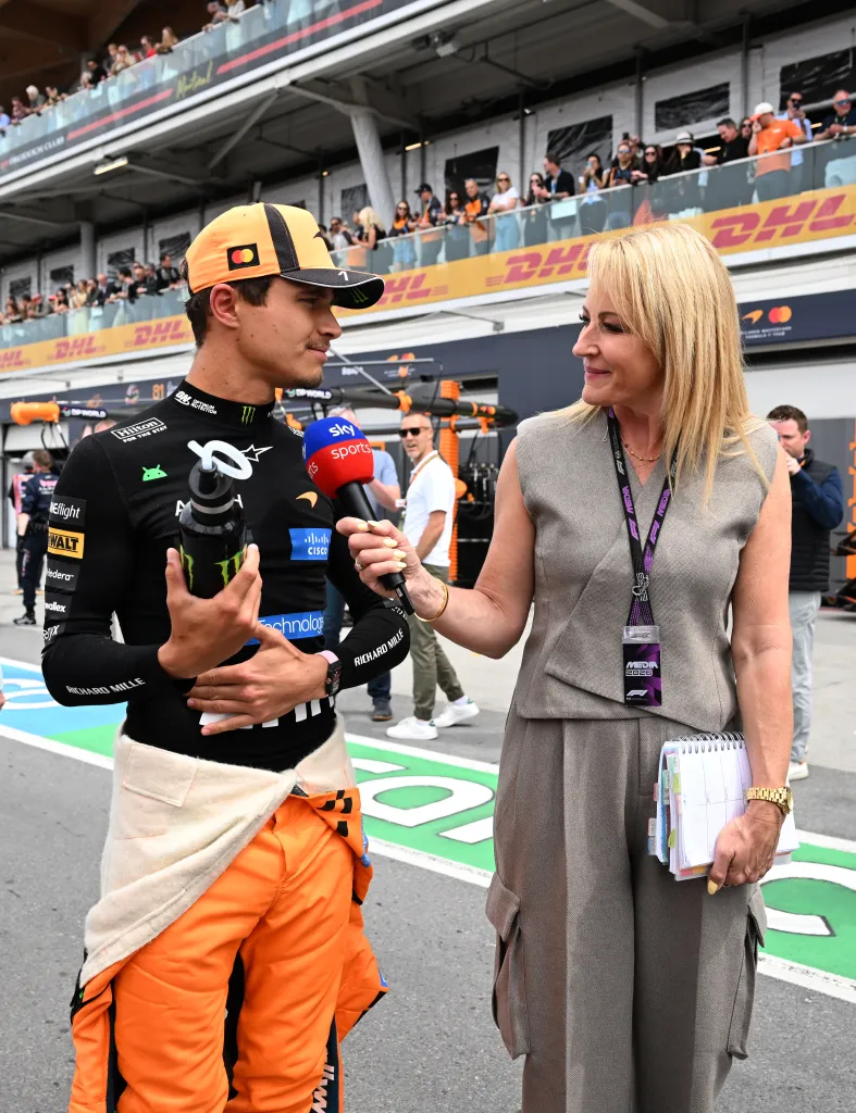 A male Formula 1 driver holding a water bottle and wearing a black racing suit and an orange McLaren hat is being interviewed by a female reporter holding a Sky Sports microphone.