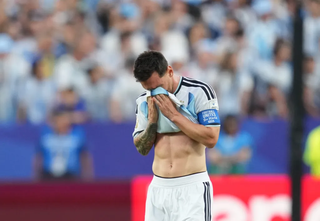 Soccer player wiping his face with his jersey, showcasing his abdomen and arm tattoos, with blurred spectators in the background.