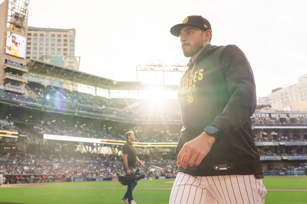 San Diego Padres pitcher Joe Musgrove walks onto a baseball field, with sun flare behind him and a stadium full of fans.