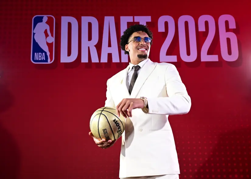Yaxel Lendeborg in a white suit, holding a golden basketball, smiling against a red backdrop with "NBA DRAFT 2026" written on it.