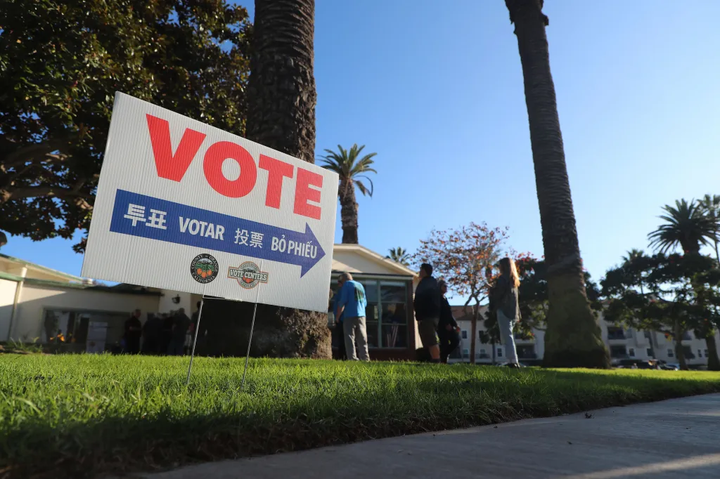 A white and blue "VOTE" sign in English, Korean, Spanish, Chinese, and Vietnamese on a grassy lawn with a building and people in the background.