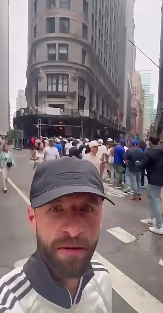 A man in a baseball cap taking a selfie with a crowd of people and buildings in the background.