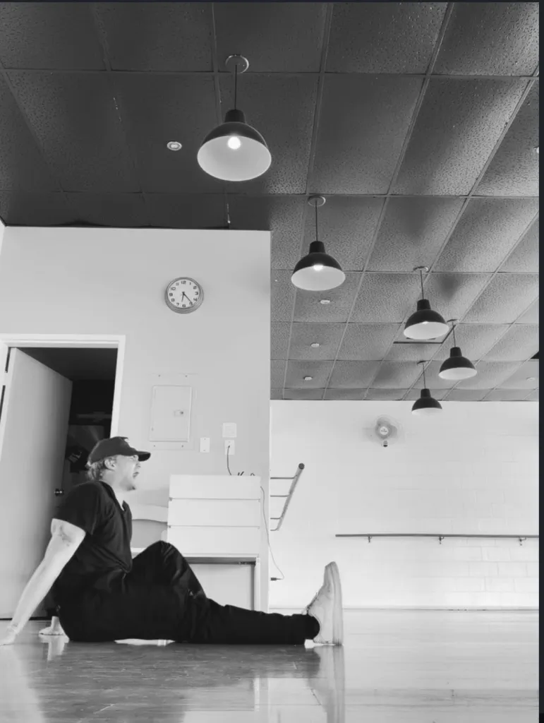 A person wearing a cap, t-shirt, and pants sits on a polished floor with one leg stretched out and the other bent, looking up in a room with industrial lights and a clock.