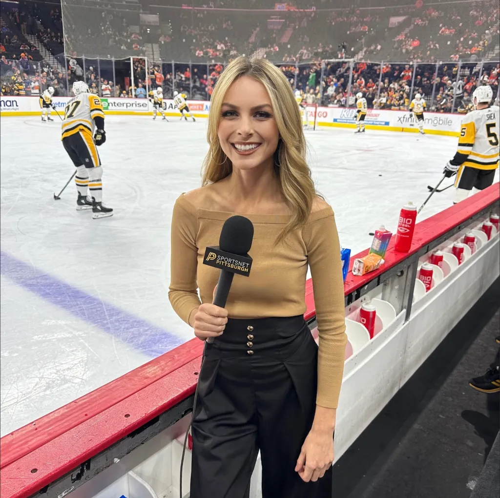 A female reporter holds a microphone with the Sportsnet Pittsburgh logo, standing on the edge of a hockey rink during a game.