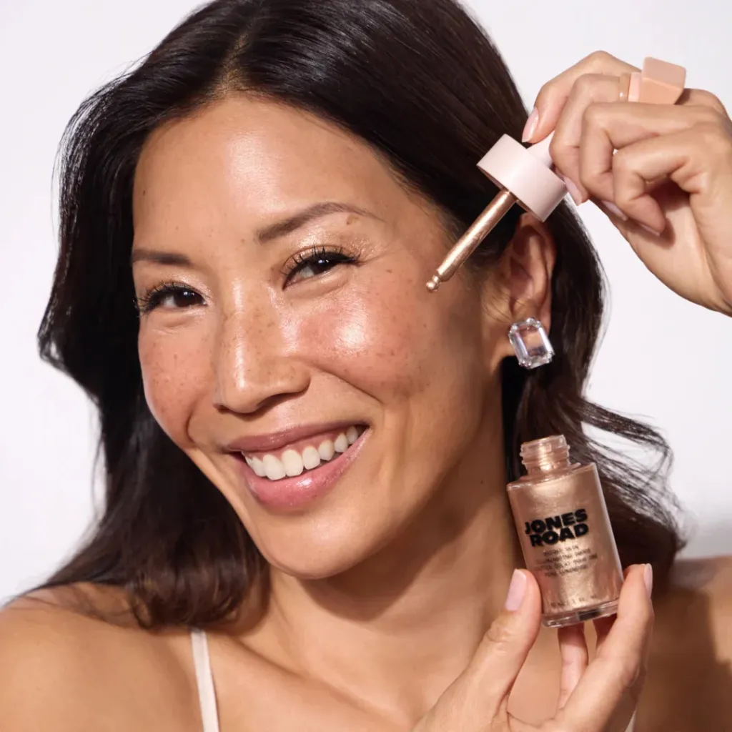 Smiling woman holding a Jones Road illuminating drops bottle and dropper.