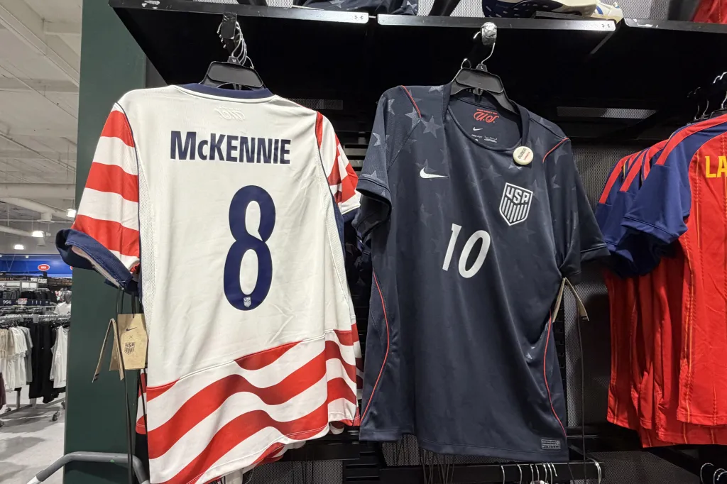 Two USA soccer jerseys, one white with red stripes on the sleeves and the number 8 with "McKENNIE" on the back, and a navy one with the number 10, hanging on a rack.