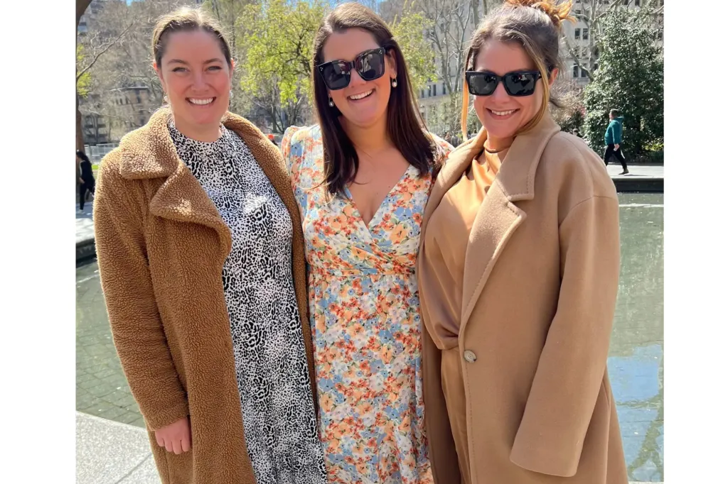 An image of three women posing together, with the one in the middle wearing a floral dress.