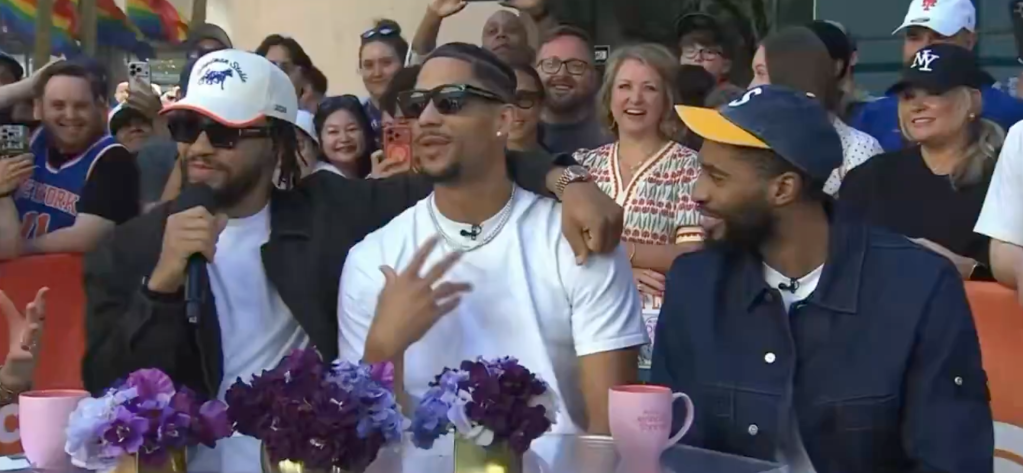 Three men are seated at a table with an audience behind them, with purple flowers and pink mugs on the table.