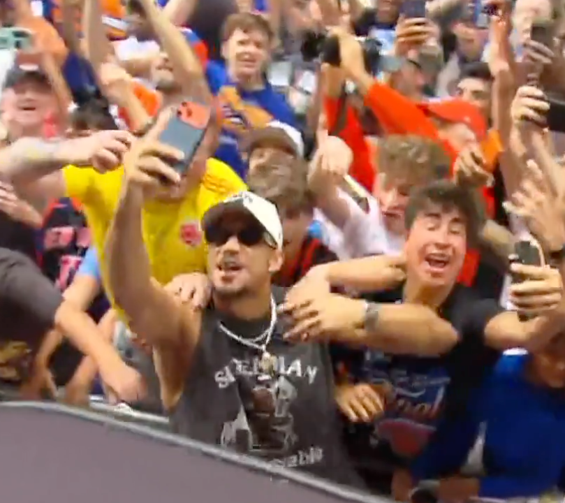 A man in sunglasses taking a selfie with a crowd of cheering people behind him.