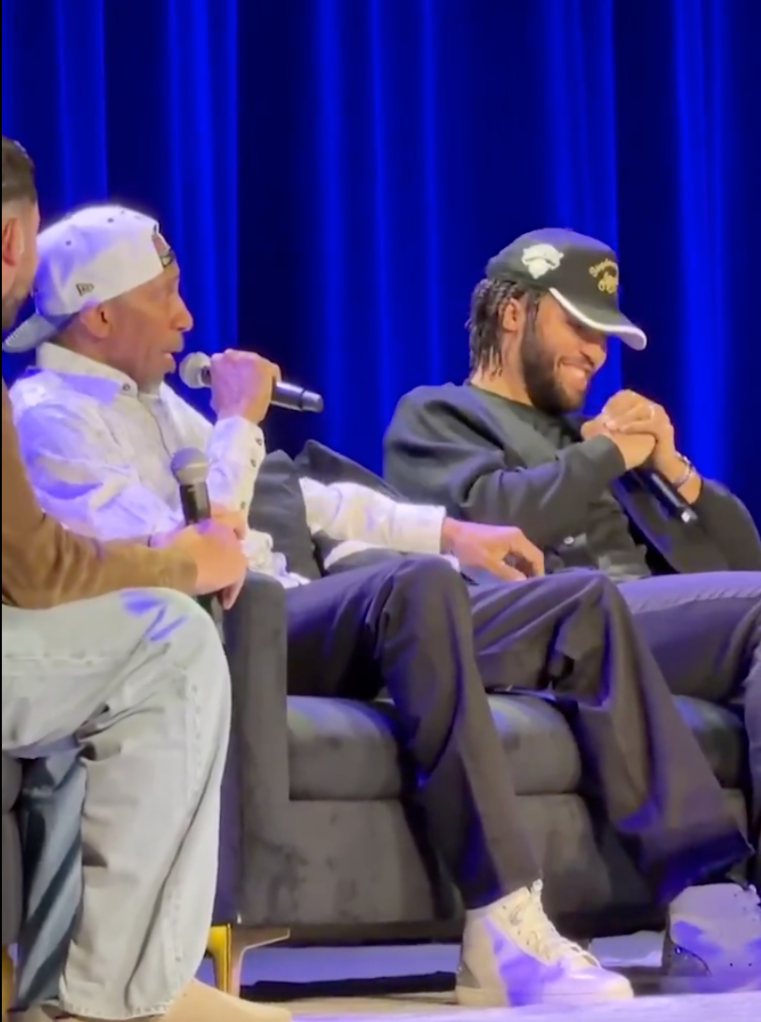 Two men in hats speaking into microphones on a couch in front of a blue curtain.