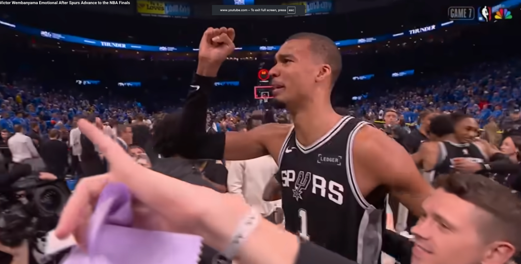 A man in a Spurs jersey raises his fist in a stadium full of people.