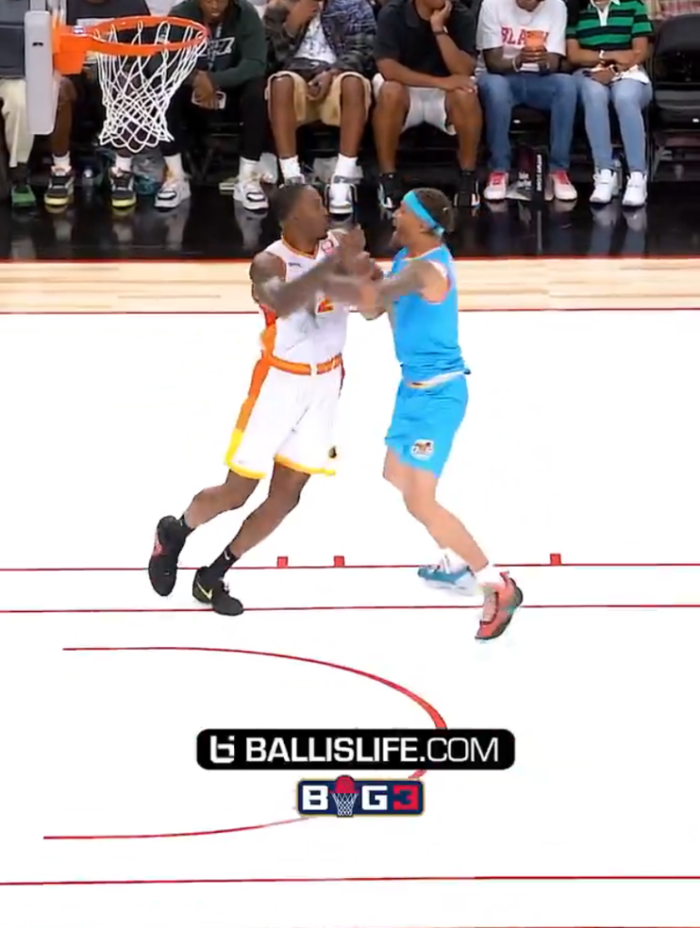 Two basketball players vying for position on the court, with a basketball hoop in the background and spectators seated behind it.