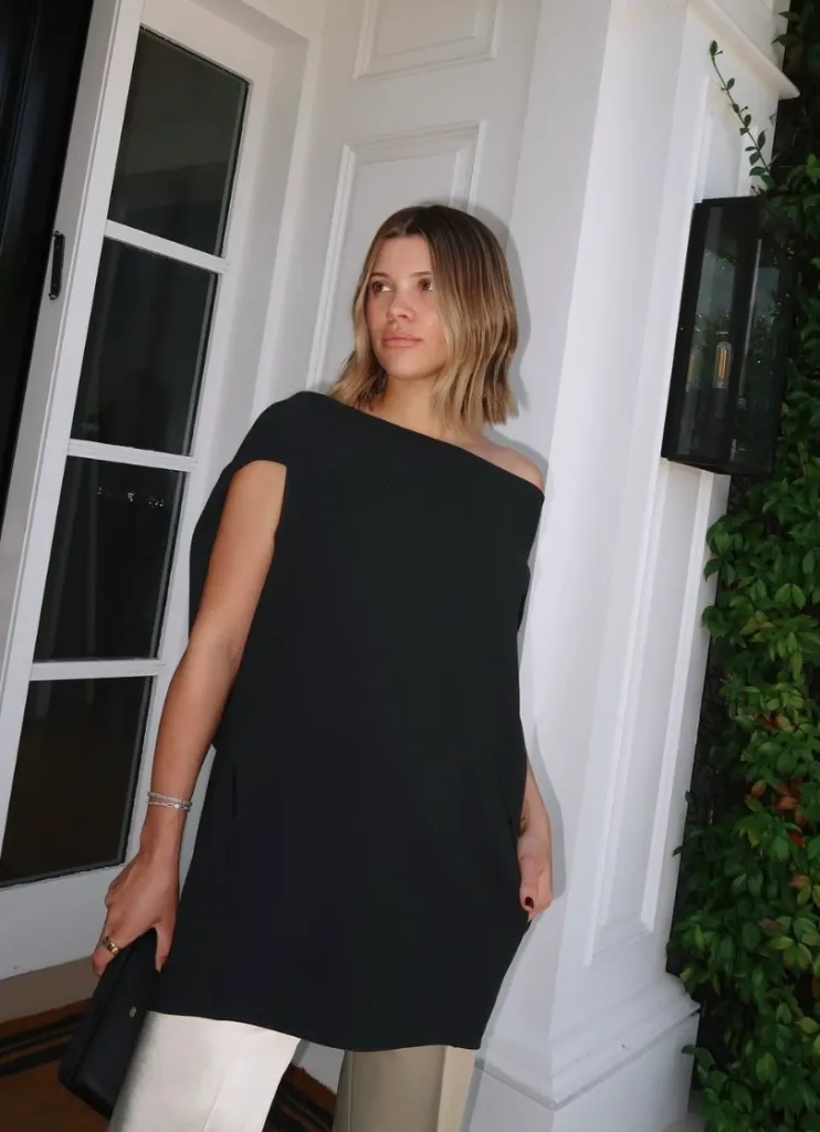 A young woman with shoulder-length blonde hair, wearing a black off-the-shoulder tunic and light-colored pants, stands near a white door, holding a black clutch.