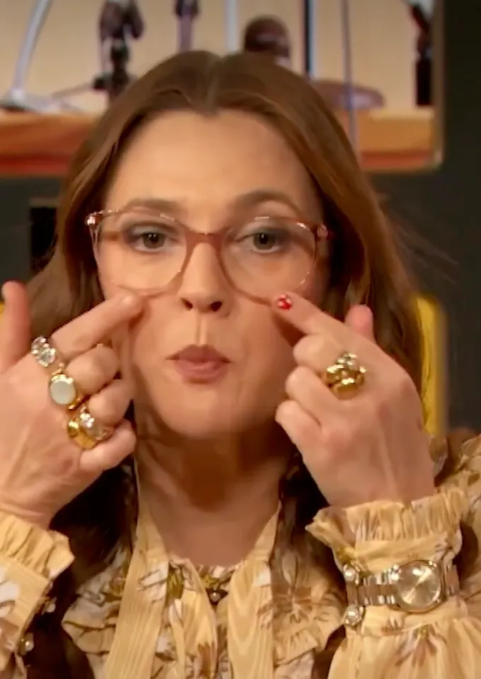 Drew Barrymore wearing glasses and rings, touching her face.