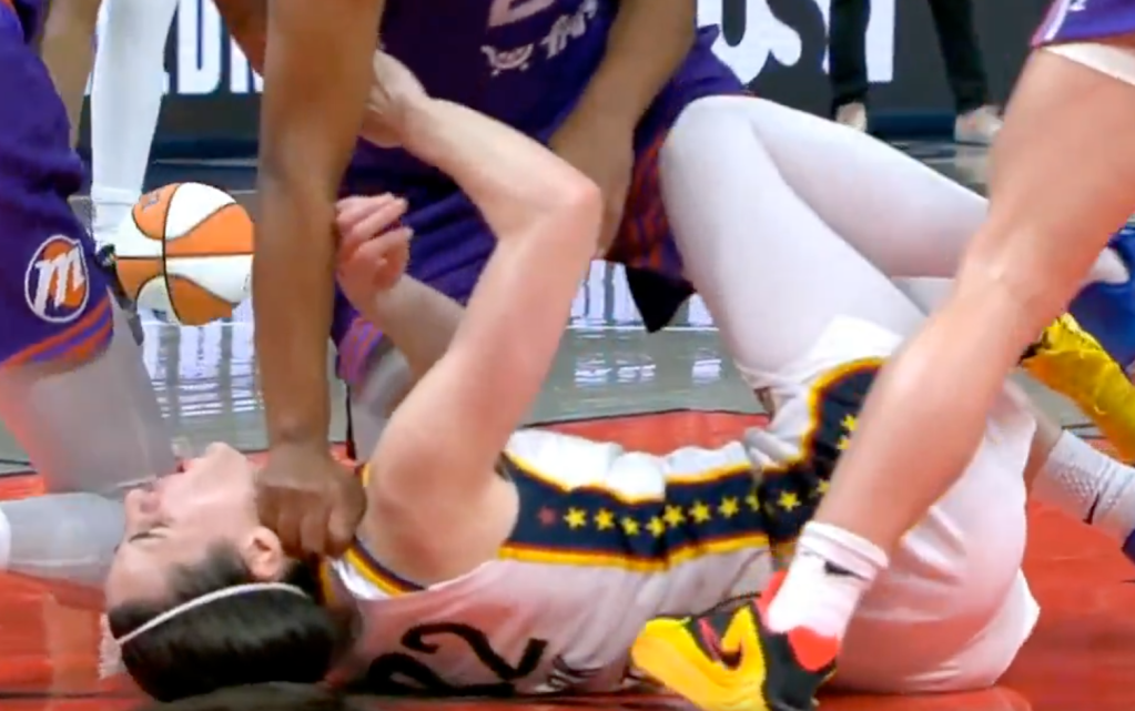 A basketball player in a white and gold uniform with stars on her shorts is on the ground while another player in a purple uniform appears to be grabbing her by the neck.