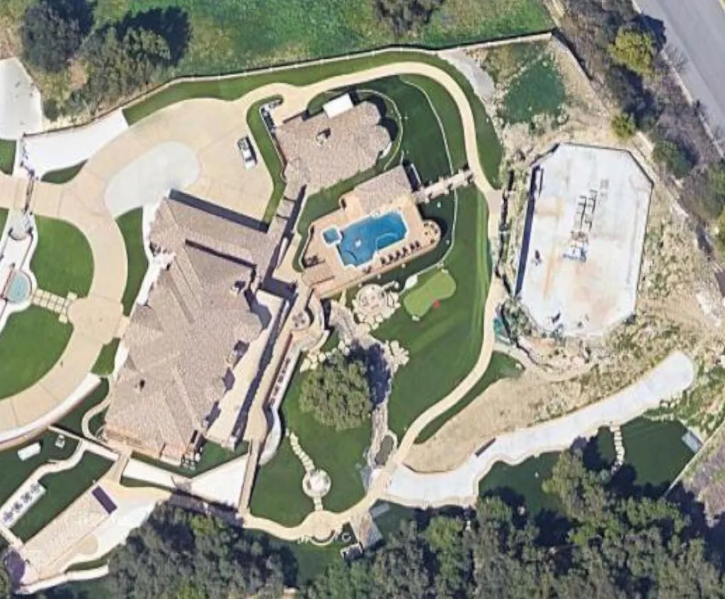 Aerial view of a sprawling property with multiple buildings, a swimming pool, a golf putting green, and a large concrete pad under construction.
