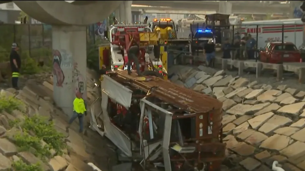 A damaged, overturned Duck Boat lies on a stone embankment next to a concrete bridge, with emergency personnel and vehicles nearby.