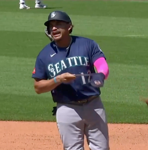 A baseball player in a Seattle Mariners jersey, with a pink sleeve, adjusting his hat.