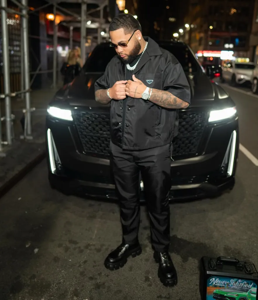 A man in a black shirt, pants, and boots stands in front of a dark car on a city street at night.
