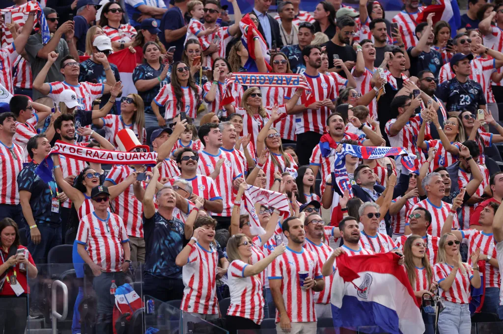 Spectators in red and white striped jerseys cheer and hold up scarves and a flag.