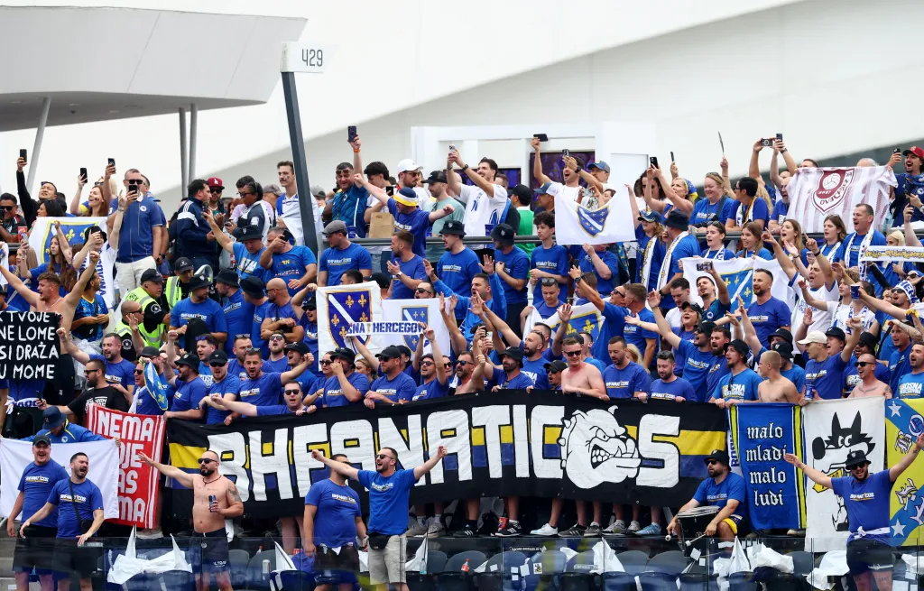 Bosnian fans in blue shirts and hats holding banners and flags, some with their arms raised, on a street before a World Cup game.