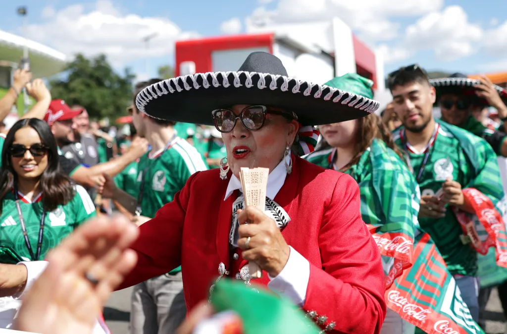 Mexican soccer fan in a sombrero and red jacket holding a fan, surrounded by other fans in green jerseys.