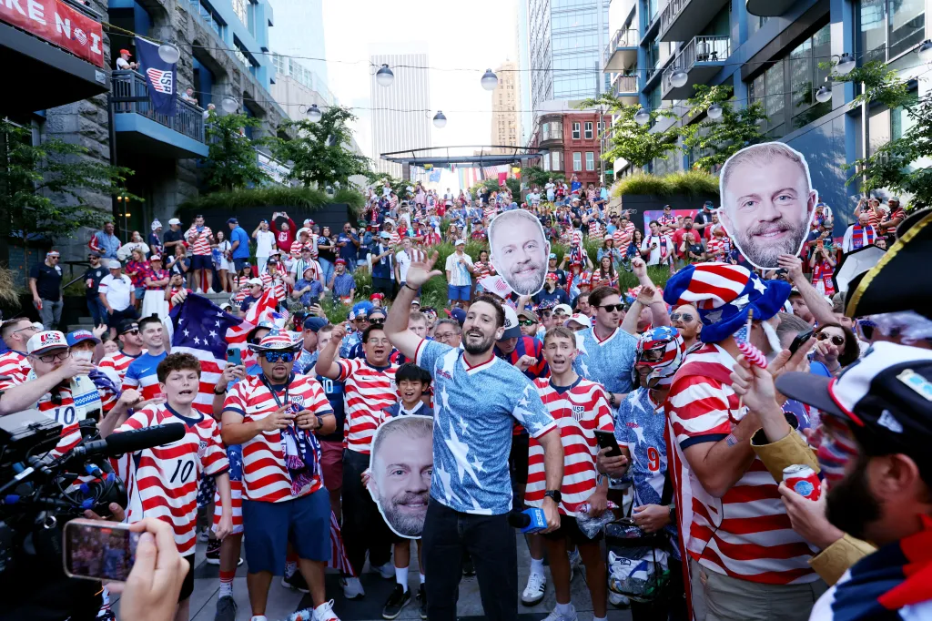 A large crowd of USA soccer fans, many wearing red, white, and blue, cheer and hold up cutouts of a man's face.