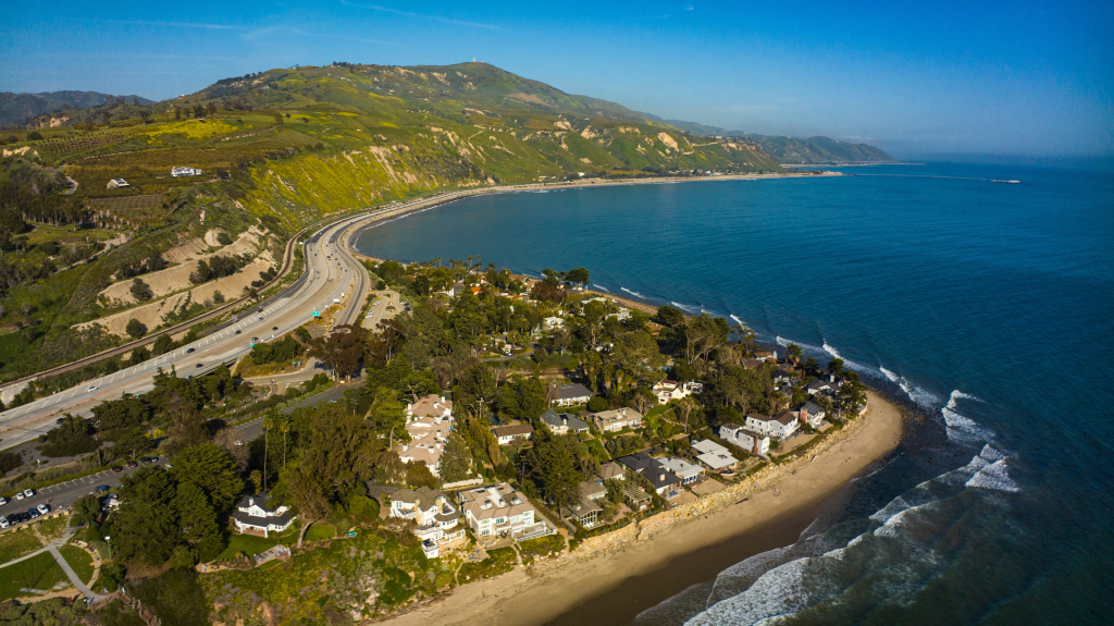 Aerial view of the California coastline with a highway running alongside the ocean, houses on the beach, and green hills in the background.
