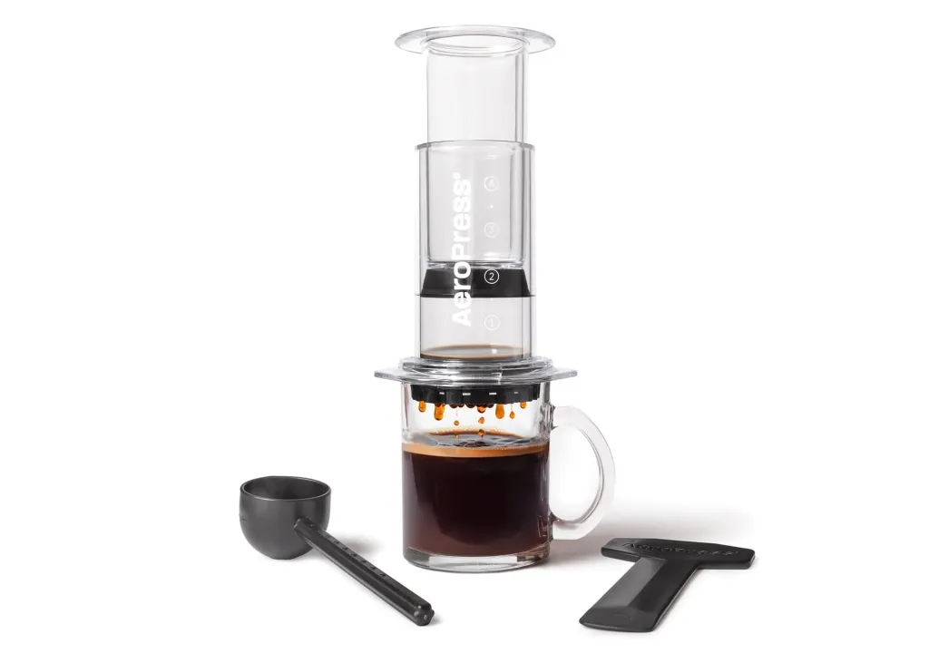 AeroPress coffee maker pressing coffee into a clear mug, with a scoop and stirrer next to it.