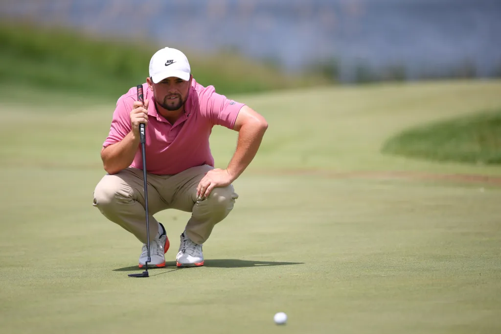 A male golfer in a pink polo shirt and a white cap crouches on a golf course, looking at the golf ball and holding a putter.