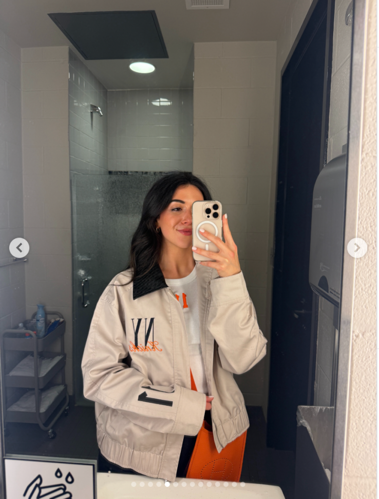 A woman takes a mirror selfie in a bathroom, wearing a beige jacket with black and orange details.