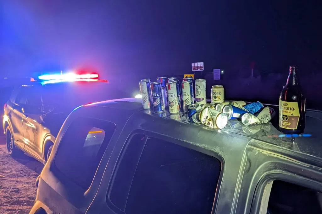 Beer bottles and cans on the roof of a car with a police car's lights flashing in the background.