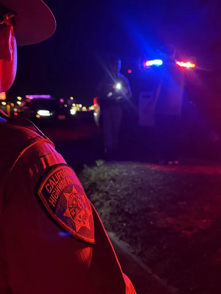A California Highway Patrol officer stands at night with police lights flashing in the background.