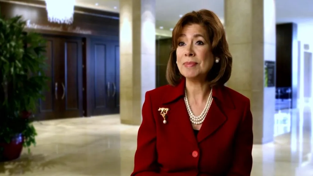 Maria Contreras-Sweet, wearing a red jacket and pearl necklace, smiles against a blurred background.